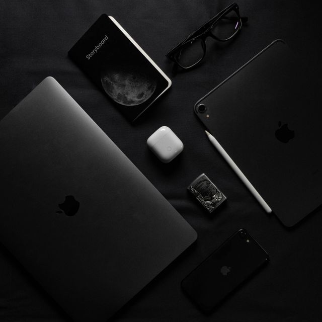 Black Apple devices, glasses, AirPods case, Apple Pencil, notebook, and a small box arranged on a dark surface